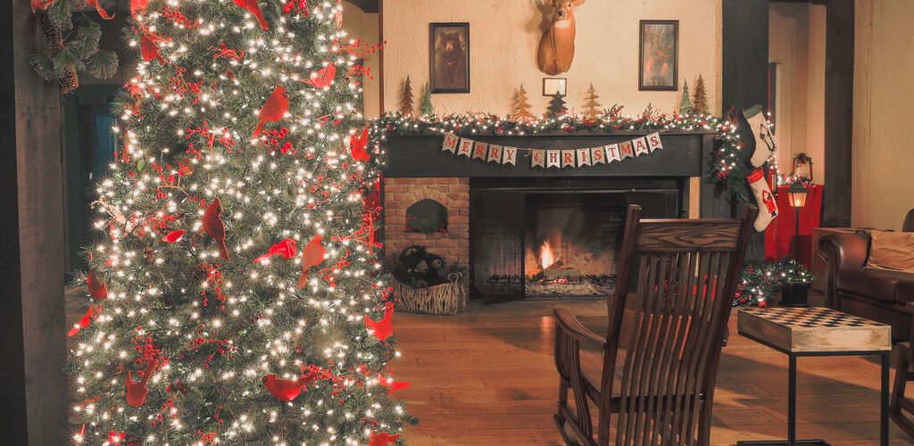 A Christmas tree in a cozy room with a fireplace. There is a taxidermied deer above the hearth and a rocking chair in front of the fireplace.  